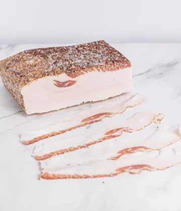 Dünn geschnittener Speck aus Colonnata, der auf einem Holzbrett mit frischen Kräutern angerichtet wird. My Little Italy