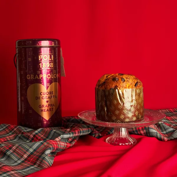 Panettone Grappa Poli Grappolone presentato su un piatto di cristallo con la sua scatola da collezione viola decorata con un cuore dorato su un festoso sfondo rosso con tessuto tartan.