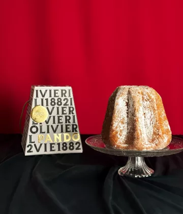 Pandoro Olivieri cosparso di zucchero a velo presentato su un piatto di cristallo con la sua scatola tipografica bianca su sfondo rosso festivo