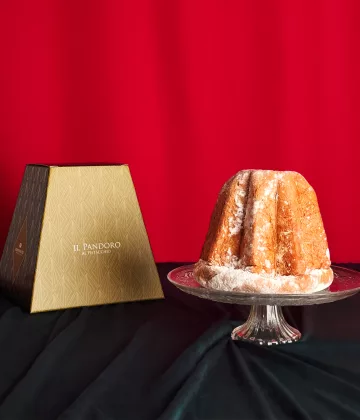 Pandoro Bonfissuto saupoudré de sucre glace présenté sur un plat en cristal avec sa boîte pyramidale beige et brune sur fond rouge festif