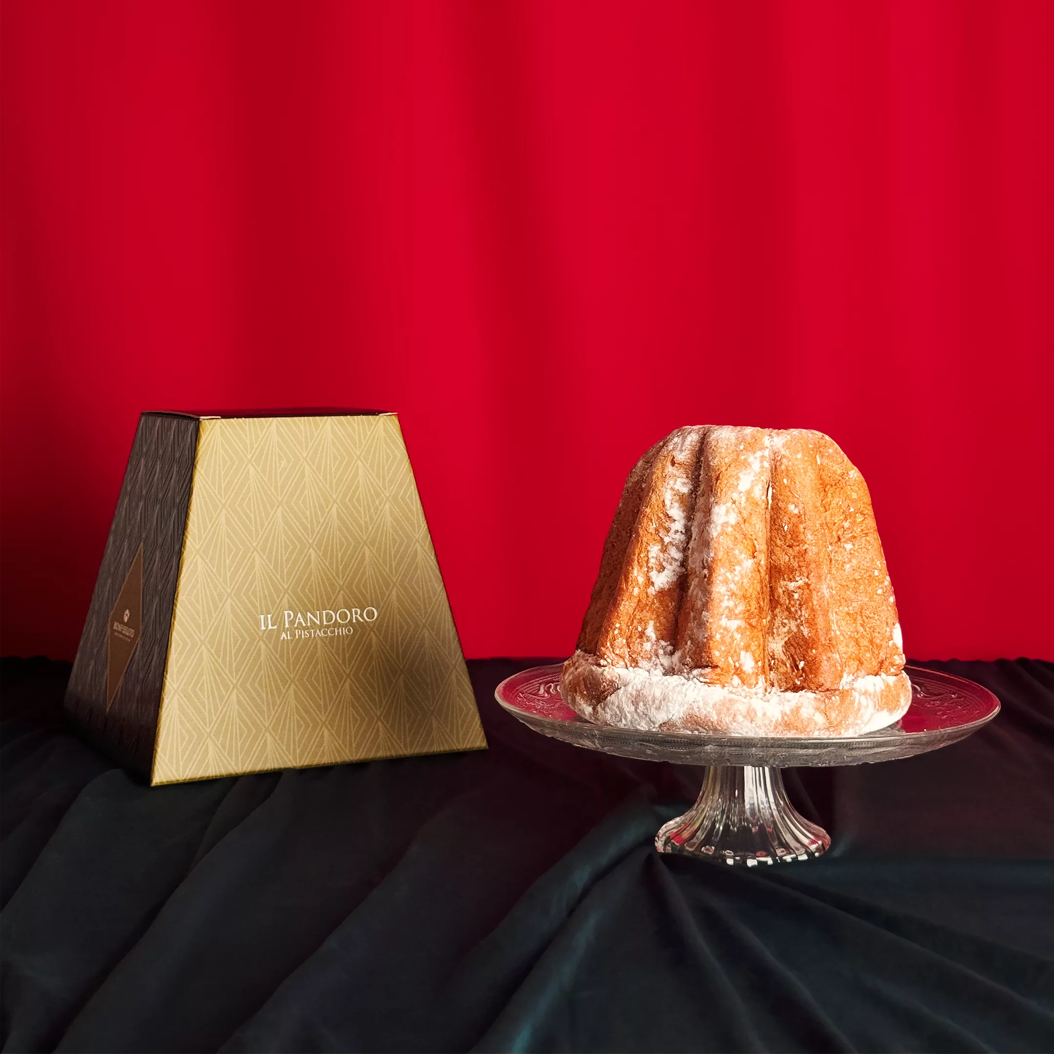 Pandoro Bonfissuto saupoudré de sucre glace présenté sur un plat en cristal avec sa boîte pyramidale beige et brune sur fond rouge festif