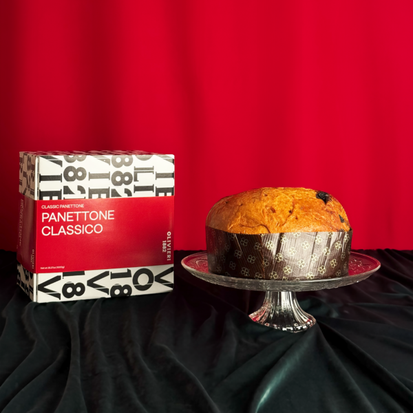 Panettone classico Olivieri presentato su un piatto di cristallo con la sua scatola tipografica rossa e bianca su sfondo rosso festivo.