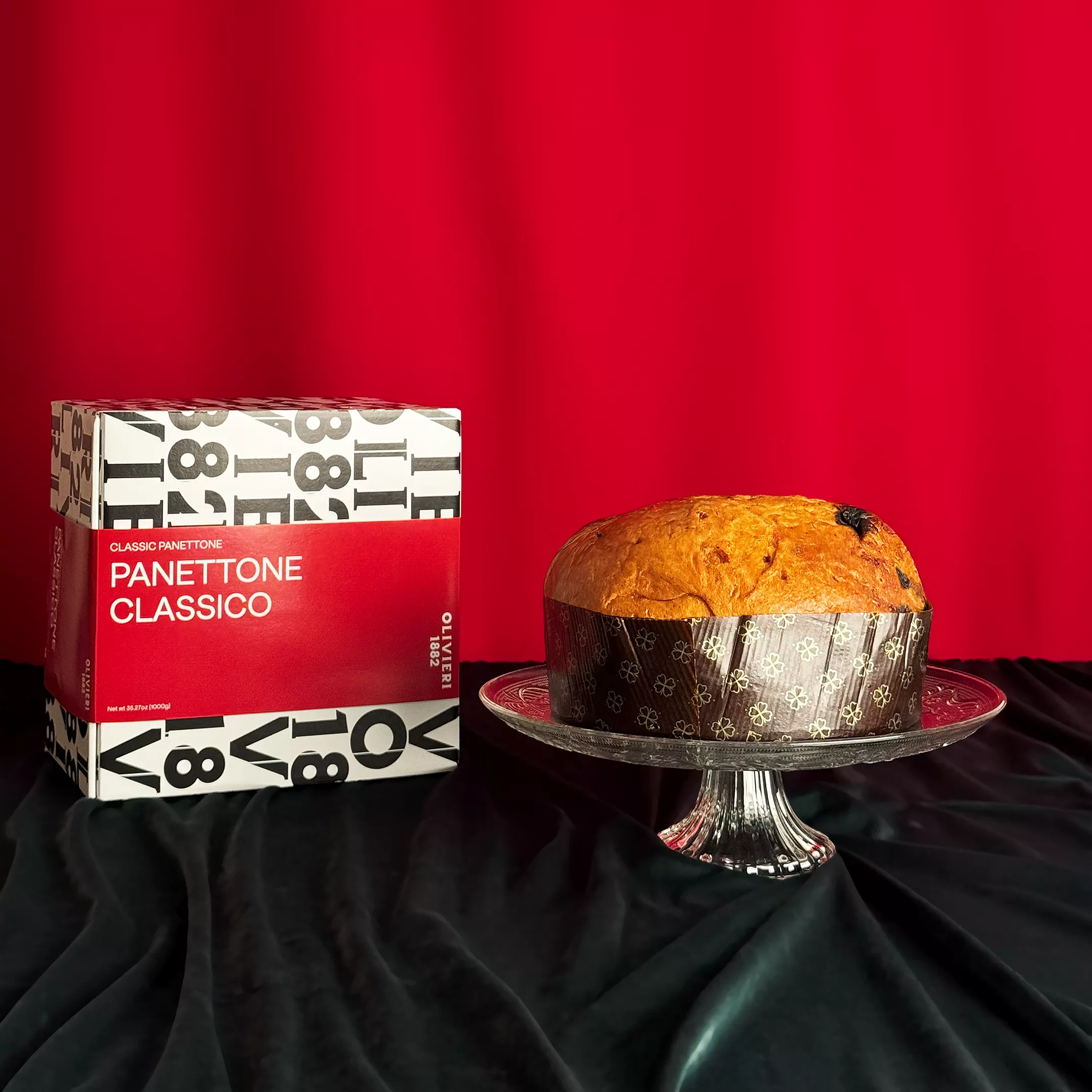 Panettone Classique Olivieri présenté sur un plat en cristal avec sa boîte rouge et blanche typographique sur fond rouge festif