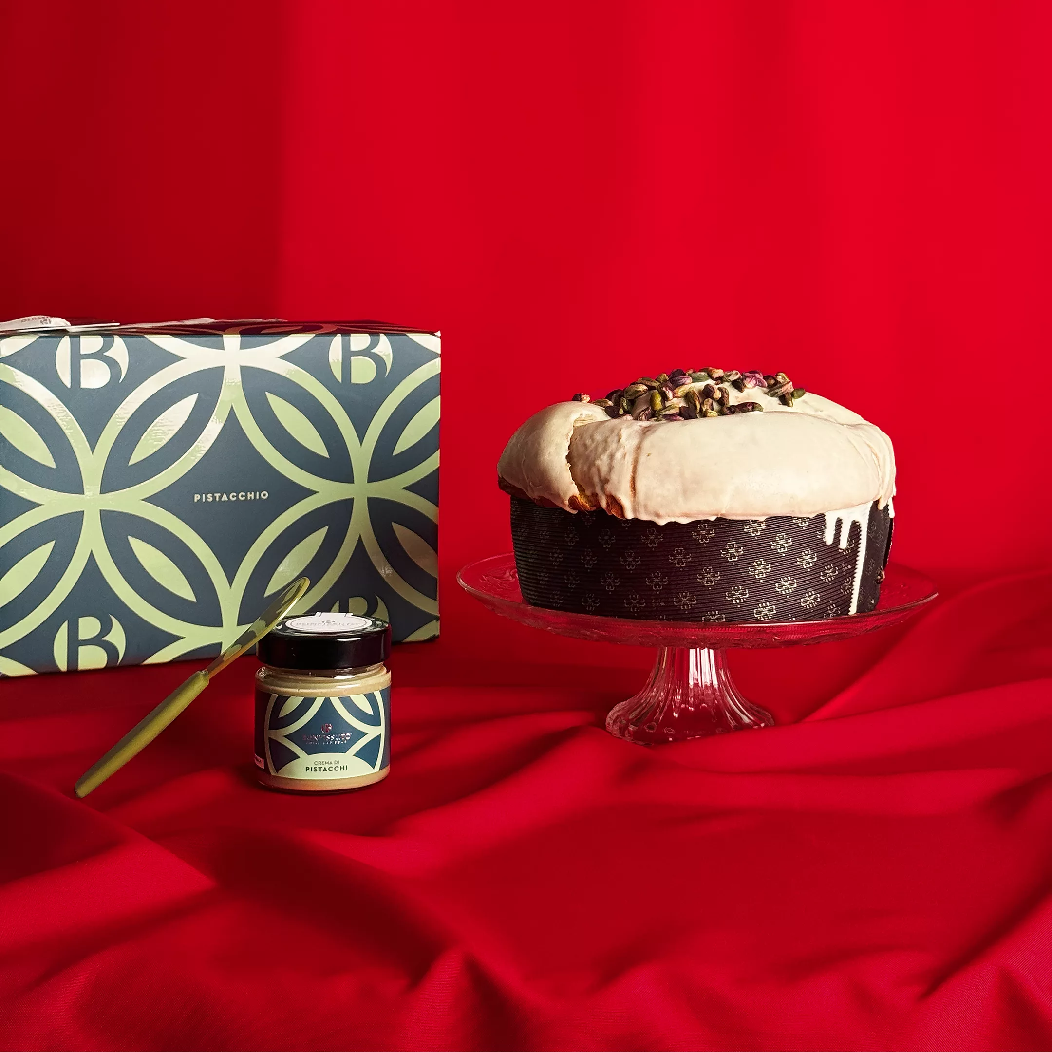 Panettone Pistache Bonfissuto avec sa couverture blanche et sa crème de pistache présenté sur un plat en cristal, accompagné de son coffret vert et de son pot de crème sur fond rouge festif