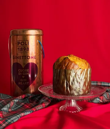 Panettone Gin Poli Ginettone auf einer Kristallplatte mit seiner kupferfarbenen Sammlerschachtel, die mit einem lila Herz auf festlich rotem Hintergrund mit schottischem Stoff verziert ist.