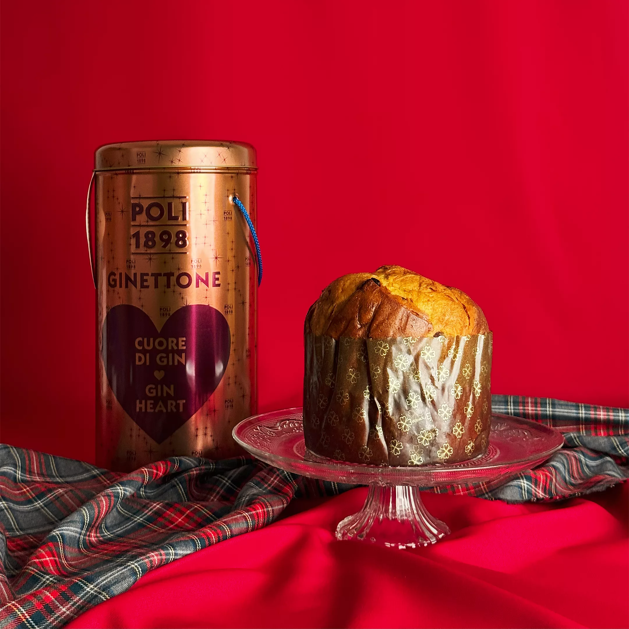 Panettone Gin Poli Ginettone présenté sur un plat en cristal avec sa boîte collector cuivrée ornée d'un cœur violet sur fond rouge festif avec tissu écossais.