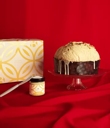 Panettone Nougat Bonfissuto avec sa couverture de chocolat blanc et son pot de crème au torrone, présenté sur un plat en cristal avec son coffret doré aux motifs géométriques sur fond rouge festif