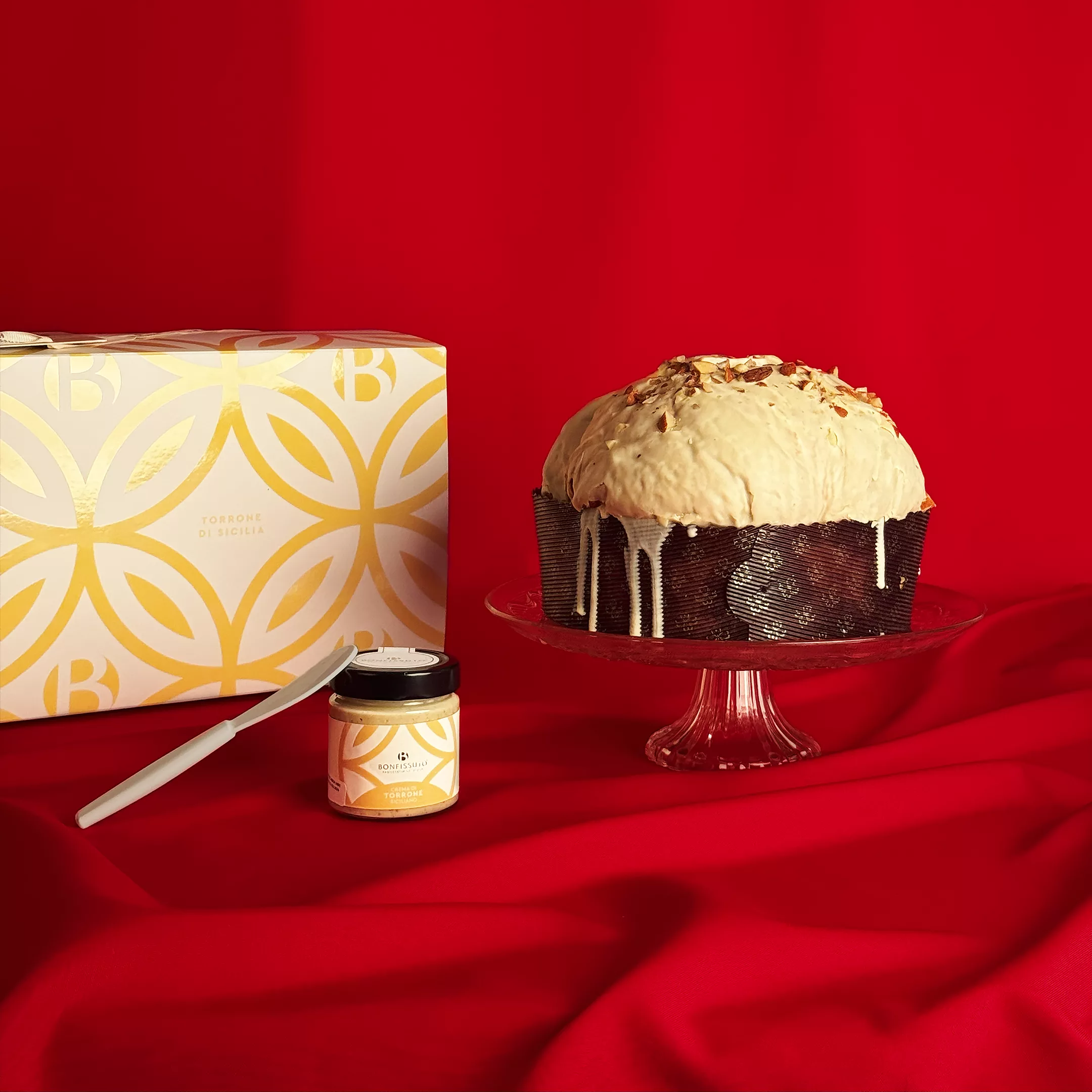 Panettone Nougat Bonfissuto avec sa couverture de chocolat blanc et son pot de crème au torrone, présenté sur un plat en cristal avec son coffret doré aux motifs géométriques sur fond rouge festif