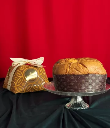 Panettone Traditionnel Bonfissuto présenté sur un plat en cristal avec son coffret doré décoré de motifs géométriques et nœud blanc sur fond rouge festif