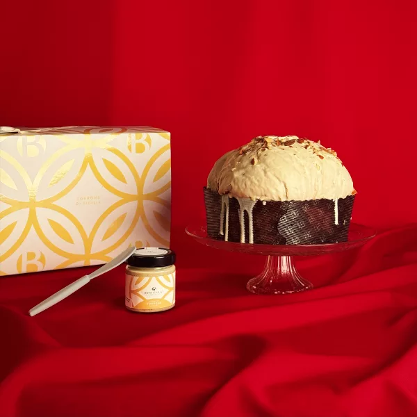Panettone Nougat Bonfissuto avec sa couverture de chocolat blanc et son pot de crème au torrone, présenté sur un plat en cristal avec son coffret doré aux motifs géométriques sur fond rouge festif