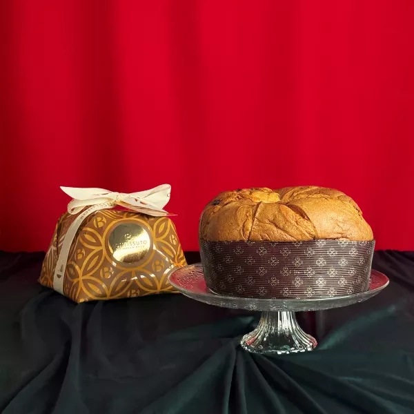Traditioneller Panettone Bonfissuto präsentiert auf einer Kristallplatte mit seiner goldenen Schatulle, die mit geometrischen Motiven und einer weißen Schleife auf festlich rotem Hintergrund verziert ist.