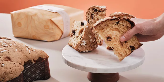 Colombe pasquali artigianali italiane: una mano che regge una fetta di torta morbida con zuccherini su un espositore di marmo, accanto a una colomba intera nella sua raffinata confezione.